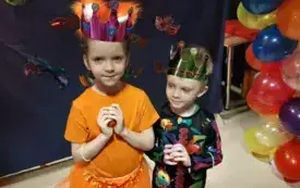 dziewczynka w pomarańczowym przebraniu i chłopiec w kolorowym przebraniu z koronami na głowach w tle balony 
