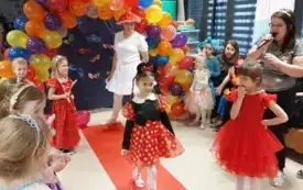 dziewczynka w tiulowym przebraniu czarno czerwono w białe kropki z opaską myszki Mini na czerwonym dywanie dookoła inne dzieci i pani nauczycielka z mikrofonem