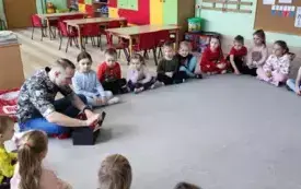 dzieci siedzą na dywanie w kole między nimi pan przed kt&oacute;rym stoi czarne pudełko do kt&oacute;rego pan zagląda
