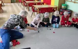 dzieci siedzą w kole na dywanie wszyscy w czepkach ochronnych pan coś prezentuje dzieciom