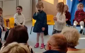 dziewczynka z mikrofonem pośr&oacute;d innych dzieci w tle dekoracja na dzień babci i dziadka
