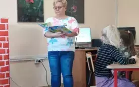 kobieta z książką w ręku w tle komputer i obrazy na ścianach widać dw&oacute;jkę dzieci przy stolikach