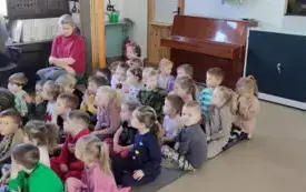 grupa dzieci na dywanie obok na krześle pani nauczycielka dzieci w coś się wpatruja