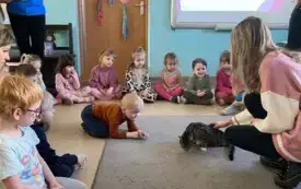 dzieci siedzą na dywanie na kt&oacute;rym jest szary kot i jego opiekunka