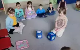 dzieci siedzą na dywanie w środku chłopiec przed nim dwie kostki jedna z kropkami druga z kolorowymi donatami, na dw&oacute;ch krzesełkach sylwety dzieci z otworami na ustach
