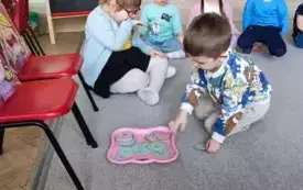 na dw&oacute;ch krzesełkach sylwety dzieci na dywanie przed nimi taca z kolorowymi papierowymi donatami kt&oacute;rym przygląda się chłopiec