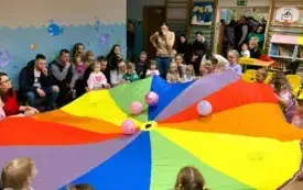 dorośli i dzieci trzymają kolorową chustę animacyjną na chuście leżą balony