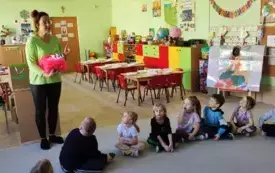 dzieci siedzą w kole na dywanie nauczycielka stoi przed dziećmi trzyma pudełko udekorowane r&oacute;żowym tiulem wypełnione upominkami