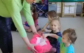 dzieci siedzą w kole nauczycielka nachyla się nad nimi z pudełkiem udekorowanym r&oacute;żowym tiulem