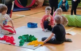 sala gimnastyczna szkolna dzieci siedzą na matach sportowych wok&oacute;ł kolorowe szarfy tunel