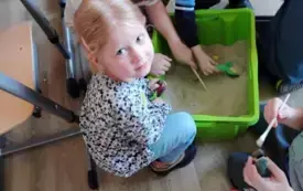 dzieci kucają dookoła plastkiowego pudełka wypełnionego piaskiem chłopiec trzyma zieloną łopatkę