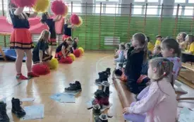 sala gimnastyczna szkolna dzieci siedzą na matach sportowych przed nimi uczennice w strojach czarno czerwonych z pomponami w rękach