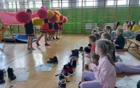 sala gimnastyczna szkolna dzieci siedzą na matach sportowych przed nimi uczennice w strojach czarno czerwonych z pomponami w rękach