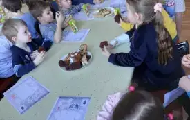 grupa dzieci siedzi przy stole przed nimi leżą pokolorowane kolorowanki oraz stoi talerz z ciastem i soczki w kartonach