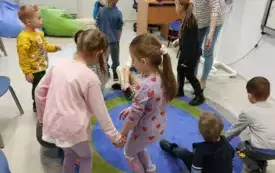 grupa dzieci siada na okrągłym dywaniku wśr&oacute;d nich nauczycielka