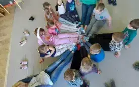 dzieci i nauczycielka na dywanie siedzą z wyprostowanymi nogami kt&oacute;re łączą sie w środku stopami w kolorowych skarpetkach