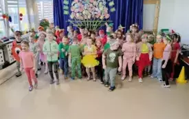 grupa dzieci w kolorowych ubraniach i opaskach owocowo warzywnych stoi na sali przed nimi rozciągnięta lina w tle dekoracja kosz z owocami i warzywami