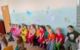 grupa dzieci w przebraniach i opaskach owocowo warzywnych siedzi na ławce pod ścianą