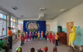 dzieci w przebraniach i opasakch owocowo-warzywnych stoją w p&oacute;łkolu po bokach nauczycielki w stroju banana i w kolorach marchewki