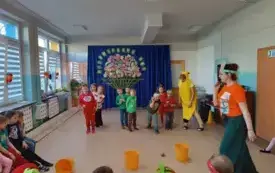 nauczycielki i dzieci w przebraniach owocowo-warzywnych na podłodze trzy kosze ż&oacute;łte, w tle dekoracja