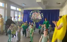 dzieci w przebraniach owocowo-warzywnych na sali między nimi nauczycielki w tle dekoracj kosz z owocami i warzywami