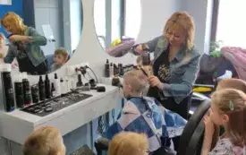 dzieci przyglądają się, pani fryzjerka czesze chłopca przed nimi lustro i r&oacute;żne kolorowe  butelki