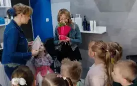 grupa dzieci wewnątrz salonu fryzjerskiego pani fryzjerka trzyma kwiata w doniczce i wącha do pani nauczycielka trzyma w ręku kartkę