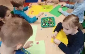 grupa dzieci siedzi przy stole na środku stoi taca z małymi pomponami dzieci trzymają pompony w dłoniach