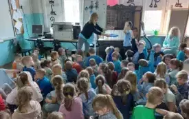 grupa dzieci w niebieskich strojach siedzi na dywanie w tle pani psycholog pani nauczycielka siedzi na krzesełku chłopiec stoi