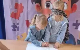 dwie dziewczynki stoją przy stoliku, jedna z dziewczynek ma oczy zawiązane chustą a druga trzyma jej dłoń na kartonie