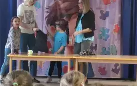 dzieci siedzą na dywanie przed nimi dwie panie i dwoje dzieci stoja przy stoliku chłopiec ma opaskę na oczach w tle dekoracja związana z dniem świadomości autyzmu