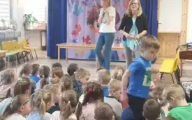 grupa dzieci siedzi na dywanie w sali gimnastycznej przed nimi dwie panie w tle dekoracja związana z dniem świadomości autyzmu