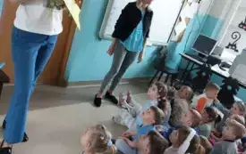 grupa dzieci w niebieskich strojach siedzi na dywanie przed nimi stoją dwie panie