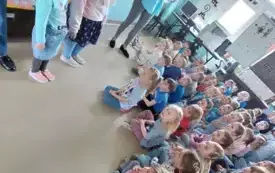 grupa dzieci w niebieskich strojach siedzi na dywanie przed nimi stoją dwie dziewczynki i pani psycholog