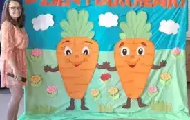 baner z napisem dzień marchewki i dwiema dużymi uśmiechniętymi marchewkami obok pani nauczycielka kt&oacute;ra na głowie ma opaskę z marchewką pozuje do zdjęcia