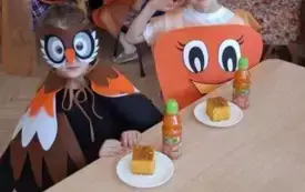 dziecko w przebraniu sowy i dziewczynka w przebraniu marchewki siedza przy stoliku przed nimi ciasto marchewkowe i soki marchwiowe w tle inne dzieci i pani nauczycielka