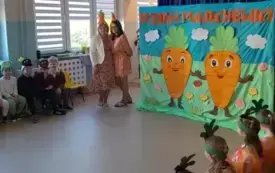 dwie panie nauczycielki w opaskach z marchewkami pozują do zdjęcia dzieci w przebraniach marchewek warzyw i zwierząt siedzą na krzesłach obok na pierwszym planie plakat z napisem dzień marchewki na kt&oacute;rym są dwie wielkie marchewki z uśmiechami