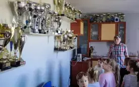dzieci ze starszym panem w pomieszczeniu w kt&oacute;rym na p&oacute;łkach stoi bardzo dużo puchar&oacute;w