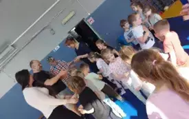 grupa dzieci wśr&oacute;d nich dwie panie i dw&oacute;ch pan&oacute;w jeden z pan&oacute;w dużo starszy, pani nauczycielka wręcza podziękowanie na piśmie panu kt&oacute;ry prowadził zajęcia