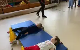 dwie dziewczynki leżą na materacu pomiędzy nogami mają pachołki obok pani nauczycielka trzyma w rękach obręcze