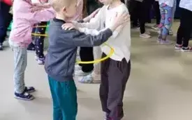 dzieci na sali stoją w parach trzymają sie za ramiona pomiędzy nimi obręcze