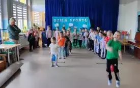dzieci stoją w dw&oacute;ch rzędach dzieci na początku trzymaja pi&oacute;lę między kolanami w tle baner z napisem dzień sportu