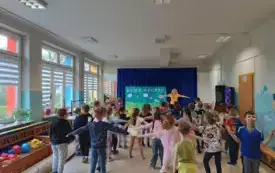 grupa dzieci stoi w rozkroku z rozłożonymi na boki rękami przed nimi pani nauczycielka w tej samej pozie widać plakat dzień sportu