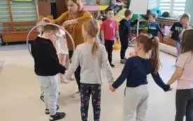 dzieci stoją w kole trzymają się za ręce w środku nauczycielka kt&oacute;ra trzyma hula hop założone na chłopca