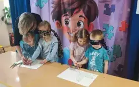 dwie pary dzieci dziewczynka prowadzi rękę dziewczynki kt&oacute;ra ma zawiązaną opaske na oczach dziewczynka prowadzi rękę chłopca kt&oacute;ry ma zawiązaną opaskę na oczach nad dziewczynkami pochylona pani w tle dekoracje związane z dniem świadomości autyzmu
