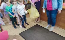dzieci stoją w rzędzie obok pani z reklam&oacute;wką chłopiec trzyma jajo styropianowe dookoła regały z książkami