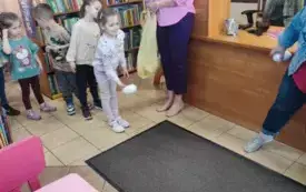 dzieci stoją w rzędzie obok pani z reklam&oacute;wką dziewczynka rzuca jajo styropianowe dookoła regały z książkami
