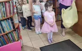 dzieci stoją w rzędzie obok paniz reklam&oacute;wką trzyma w dłoni jajo styropianowe dookoła regały z książkami