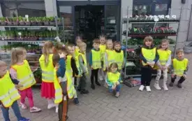grupa dzieci w kamizelkach odblaskowych pozuje do zdjęcia przed wejściem do sklepu ogrodniczego w tle wejście do sklepu i regały z sadzonkami