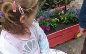 dziewczynka kuca przy wagoniku ze skrzynki w kt&oacute;rym posadzone są kolorowe kwiaty